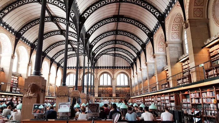 Photo 1 of Biblioteque Genevieve interior, a large room with very high vaulted ceilings and arched windows. The walls are lined with bookshelves and there are desks all filled with people in the centre.