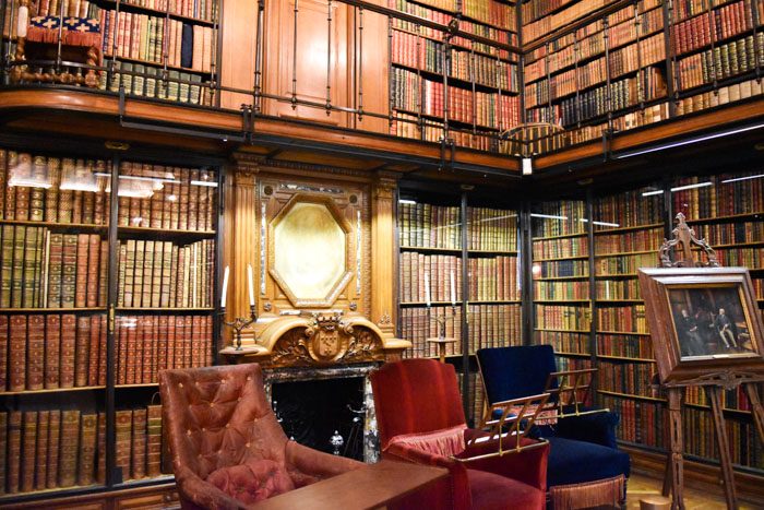 Photo of Chantilly Castle library interior 2, a different angle of the same room, showing bookshelves in the background, an ornate fireplace in the center, and three padded chairs and a painting on a stand in the foreground.