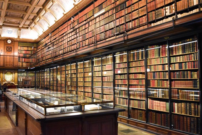 Photo of Chantilly Castle library interior, a large room with all the walls covered in bookshelves, including an upper level with a balcony rail. In the center of the room a a glass display case with more books inside.