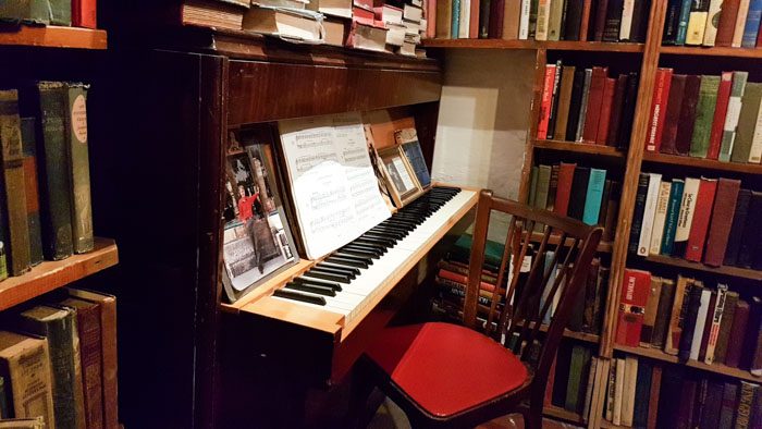 Photo of the interior of Shakespeare & Company, an upright piano surrounded by books, a wooden chair with a red seat sits at it.