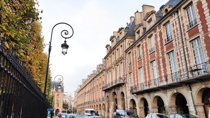 Photo of the street the Victor Hugo Museum is on. On the left are tall trees, a black metal fence and a fancy lamp post. On the right is a long row of red and white brick buildings with arches on the ground level and small decorative balconies in front of the upper windows.