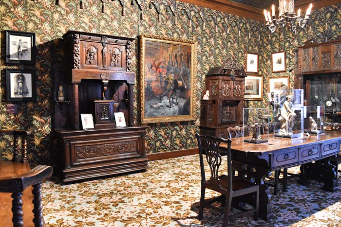 Photo of the interior of the Victor Hugo museum, showing a room with dark floral patterned wallpaper, heavy dark wood furniture, 7 paintings on the wall in frames and a large dining table which has glass display boxes on top of it.