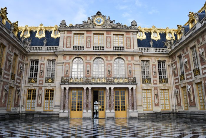 The front of the Palace of Versailles, a huge 3 storey building with a black and white patterned tile square, gilt decorations on the windows, balconies and windows. A huge clock dominates the center top, flanked by sculptures.