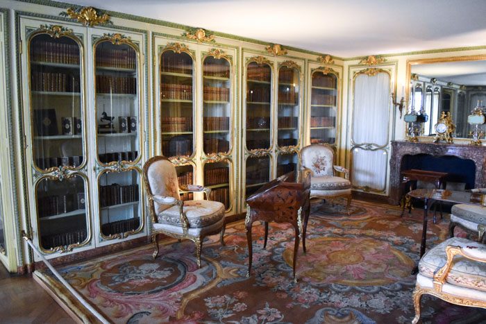 Photo of the interior of Versailles, a small room with a wall of bookcases on the left with green and gold decorative borders around the glass doors. On the right a mirror hangs above a fireplace. There is a beautiful pink and red rug on the floor and 4 padded armchairs with floral designs and a small wooden writing desk.