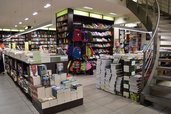 Inside Ianos Aristotelous. Bookshleves and tables stacked with many books and a curved staircase on the right.