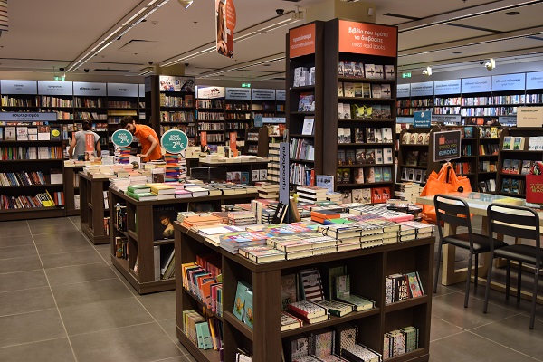 Inside of Public in Cosmos. Many dark wooden bookshelves and tales piled with books.