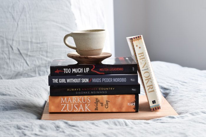 A stack of books on a bed. Bridge of Clay by Marcus Zusak, Always Another Country by Sisonke Msimang, The Girl Without Skin by Mads Peder Nordbo, Too Much Lip by Melissa Lucashenko, Starbook by Ben Okri