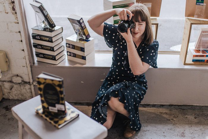 Tamsien West - Babbling Books, taking a photo in a bookstore, wearing a navy blue dress with white polka dots.