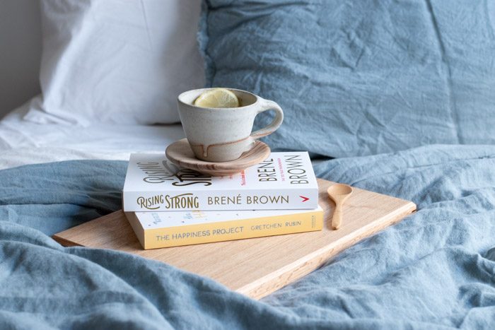 4 Ways to care for yourself as a creative. Images shows a closeup of a bed with blue and white linen. Two books are stacked with a tea cup on top. The book titles are The Happiness Project by Grechin Rubin and Rising Strong by Brene Brown.