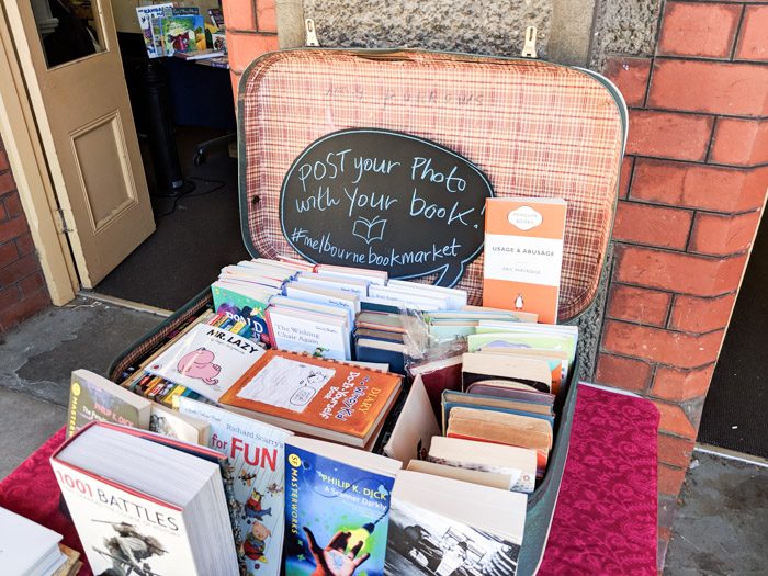 Melbourne Book Market at the Abbotsford Convent, an assortment of bocks stacked inside a vintage suitcase. A blackboard shaped like a speech bubble reads 'Post your photo with your book! #melbournebookmarket'