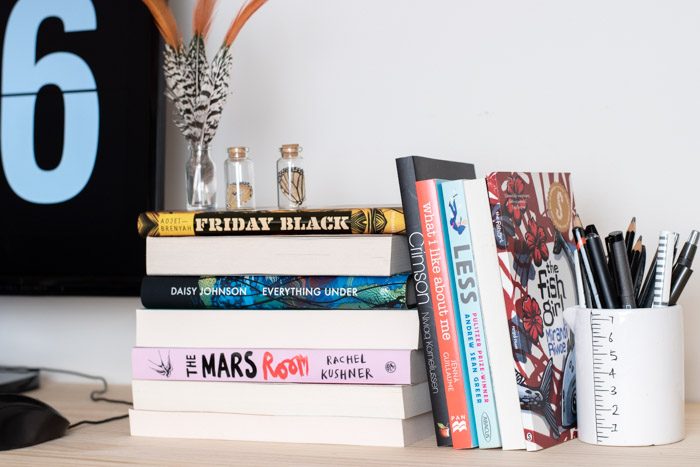 7 Books to Read from Sydney Writers Festival. Books stacked on a desk with a ceramic measuring cup used as a pen-holder. 