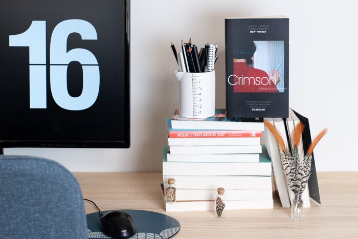 Seven Book to Read from Sydney Writers Festival. Stack of books on a desk, Crimson by Niviaq Korneliussen with a black cover and a person in a red shirt holding a cigarette is the focal point of the photo.