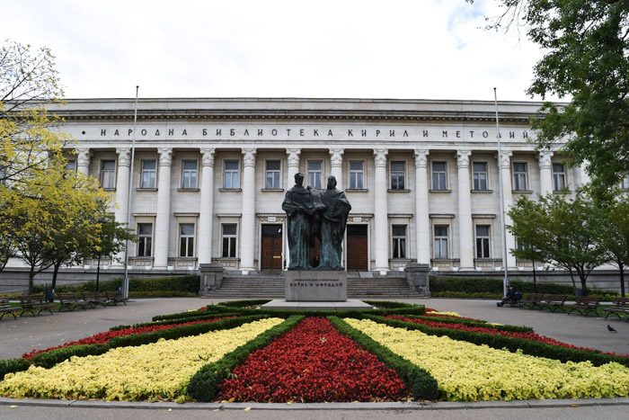 Front of the St. Cyril and Methodius National Library in Sofia, Bulgaria. An Impressive white two storey building with columns all the way along the facade. A bronze statue of two robes figures stands in front of the building, in the middle of a flowerbed of red, yellow and green geometric design.