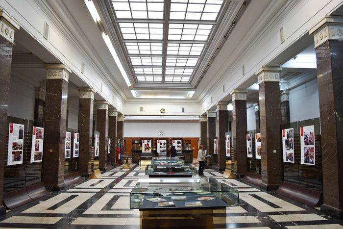 Interior of the St. Cyril and Methodius National Library in Sofia, Bulgaria. A large skylight runs the length of the ceiling. Marble columns stand on each side, and glass display cases are along the center of the room. The floor tiles are a black and white geometric design.