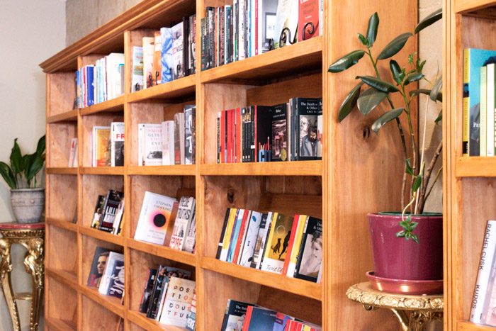 Interior of Dr B's Bookstore, Trentham. A close up photo of one of the wooden bookshelves, There are two plants on narrow gilt tables on either end of the bookshelf.