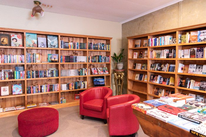 Interior of Dr B's Bookstore, Trentham. Wooden bookshelves are along two walls, and the edge of a table covered in books is just visible on the right. The shelves are sparsely  stacked with books, many books are facing forward, and two shelves are empty. There are two red tub chairs and a round red footstool in the centre of the room. Oddly, there is a sculpture of a chicken and 4 eggs on the ceiling. 