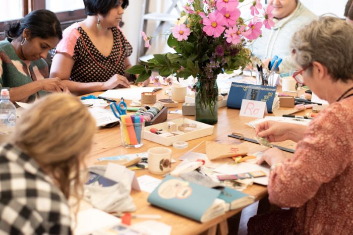 Creative Journaling Workshop Melbourne. A table filled with stamps, paper and supplies and five people are seated at the table working on creative journals.