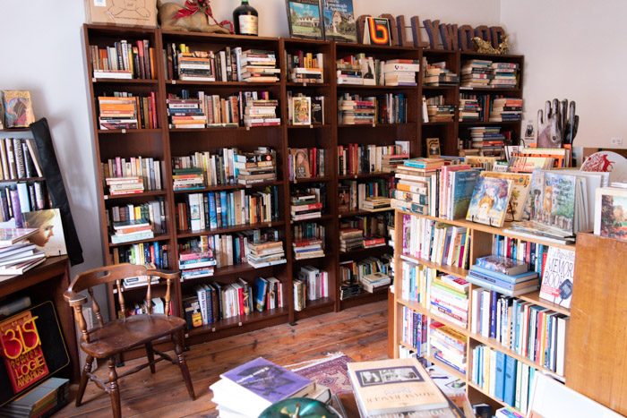 Interior of Long Story Short Secondhand Books and Music. A cosy room has a large dark wooden bookshelf on the far wall, filled with books. In the forground as a wooden chair and a smaller pale wooden bookshelf, also filled with books.