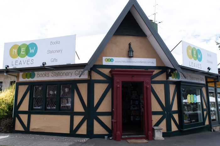 Exterior of New Leaves, Woodend. A faux Tudor style building, yellow with dark green trim and a pointed roof above the front door. 5 signs say New Leaves, Books Stationery, Gifts. 