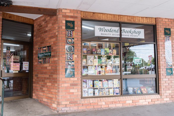 Exterior of the Woodend Bookshop, Woodend. A simple orange brick facade has a large window through which a display of books can be seen.  Multiple signs say 'WB Books' and a white sign in the window reads 'Woodend Bookshop, Quality secondhand books'.