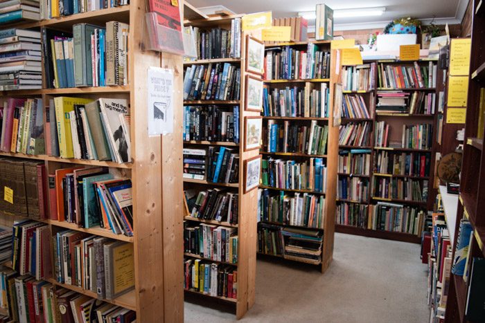 Interior of the Woodend Bookshop. Four rows of bookshelves can be seen, all stacked with books. There are bright yellow signs at the top of each shelf indicate the genres or topic of the books.