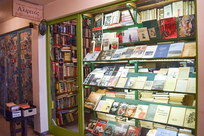 Front of Alfios Bookstore, Athens, Greece. A large window occupies most of the space, along with a glass door. Both have a bright green painted frame. books fill every space visible through the window and the door.