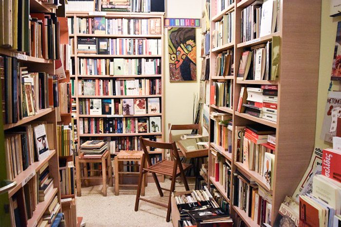 Inside Alfios Bookstore, Athens, Greece.  Colourful books are neatly stacked on wooden bookshelves. Two stools and a couple of chairs complete the scene, and an abstract artwork in brown and green hangs on the wall.