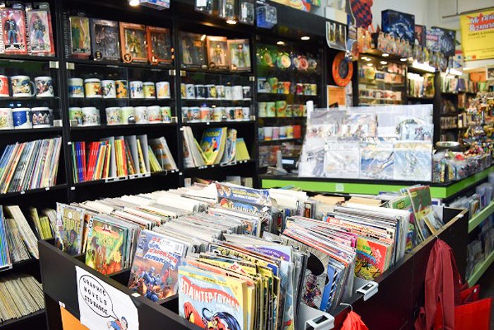 Inside Comicon, Athens, Greece. Comic books are neatly stacked in the forground and on bookshelves. Various figurines and mugs with comic book character designs can be seen on the shelves too.