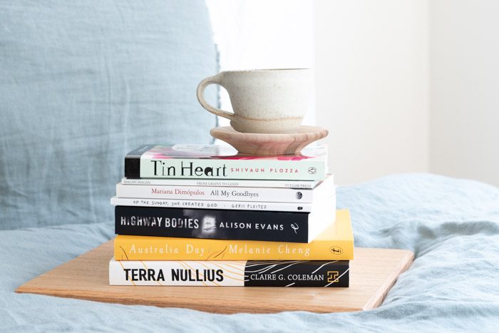 Stack of books from Emerging Writer's Festival 2019, on a bed with pale blue sheets. The titles from top to bottom are: Tin Heart, From Grains to Gold, All My Goodbyes, On The Sunday She Created God, Highway Bodies, Australia Day, Terra Nullius.