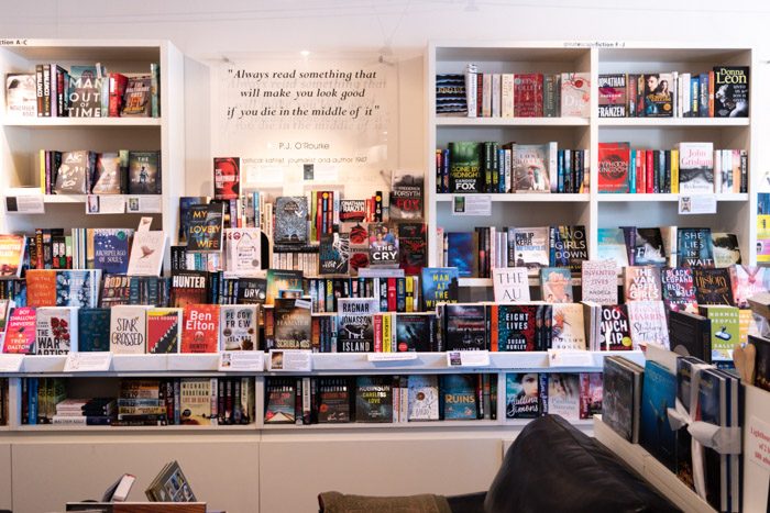 Picture of the inside of Great Escape Books, showing a huge range of books on white shelves. There's also a sign with a quote on it that reads "Always read something that will make you look good if you die in the middle of it" by PJ O'Rourke.