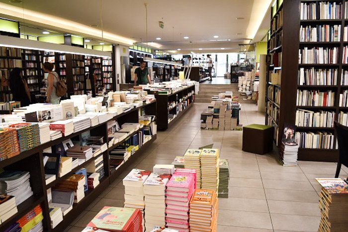 Inside Ianos Art, Athens, Greece. The walls are lined with tall dark bookshleves filled with books. The center of the long room has tables completely covered with stacks of books. There are also stacks of books on the floor.