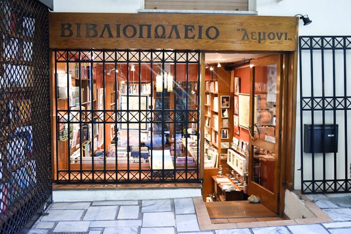 Front of Lemoni, Athens, Greece. A small shop front with a wooden sign above the window and door. The window is barred with black iron bars. Steps are visible leading down into the shop which looks warm and cosy.