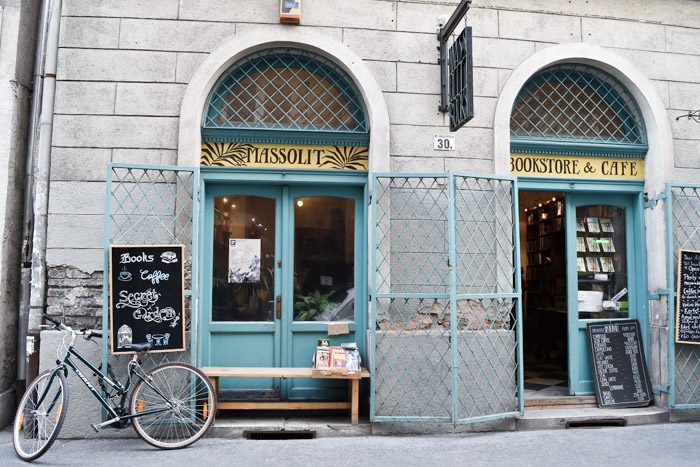 Two sets of blue arched double doors with handpainted signs above them that say 'Massolit Bookstore & Cafe', alongside several chalkboards and one bicycle.