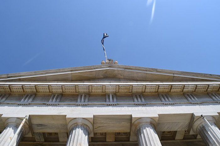 Front of the National Library of Greece, as seen from the staircase looking up. A Greek flag is waving against a blue sky.