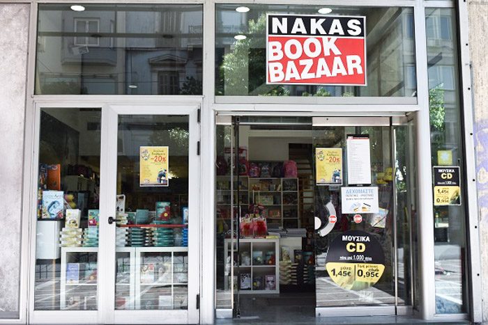 Exterior of Nakas Book Bazaar, Athens, Greece. A modern glass-fronted building. The only defining feature is a large black, white and red sign that reads 'Nakas Book Bazaar'. 