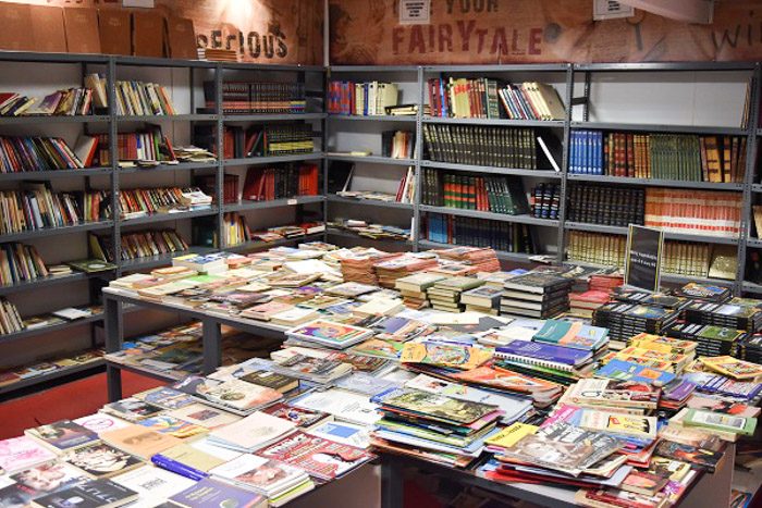 Inside Nakas Book Bazaar, Athens, Greece. Industrial style metal bookshelves line the walls and have an assortment of books on them. Some shelves are empty. The centre of the room is occupied by a table covered in books.