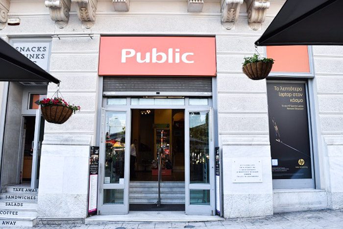 Front of Public, Athens, Greece. An imposing pale stone building with a large rectangular orange 'Public' sign above the open door.