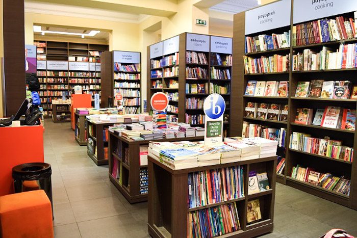 Inside Public, Athens, Greece. Wooden bookshelves line the walls and low bookshelves are evenly spaced along the center of the room. Above each shelf is a sign in Greek and English indicating the genre.