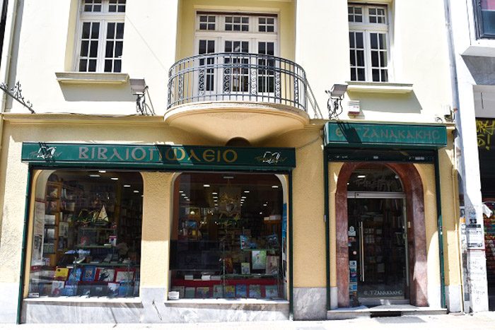 Front of Bookstore E. Tzanakakis Parimin, Athen, Greece. A pale yellow building with dark green signs above the two windows and arched front door. Books can be seen through the windows.