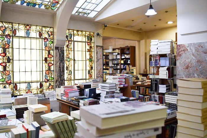 Inside Bookstore E. Tzanakakis Parimin, Athen, Greece. Piles of books fill the foreground. The room has large windows decorated with stained glass in the design of grape vines.