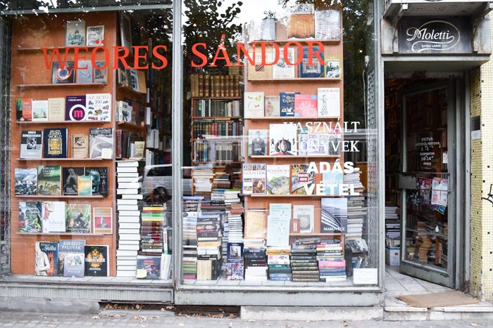 Picture of the front of this bookshop, which is floor to ceiling windows and filled with bookshelves and book piles.