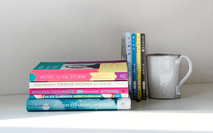 A book stack of five books, lying on their side spines facing towards the viewer, next to Japanese CDs and a coffee mug.