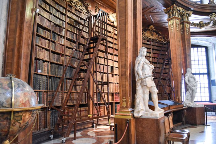 Close up of one of the large wooden bookshelves in the Austrian National Library, with a rolling staircase and two marble statues alongside. 