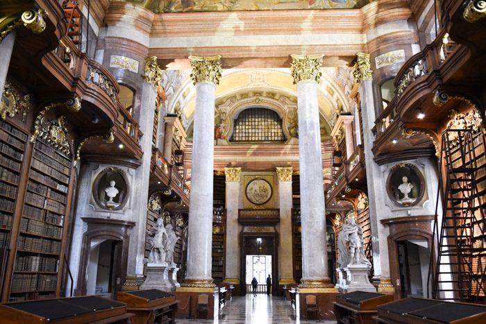 A grand hall with towering marble columns flanked by bookshelf-covered walls. 