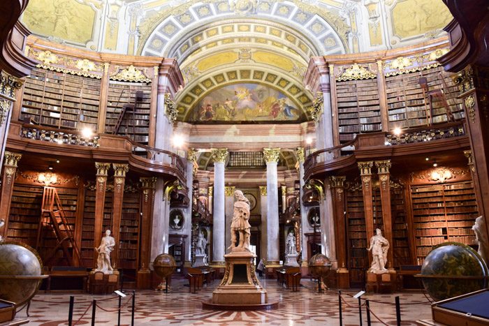 The interior of the Austrian National Library. Marble statue in the centre of the circular room, surrounded by large timber bookshelves, marble columns, and Venetian globes, topped with a domed, painted ceiling.