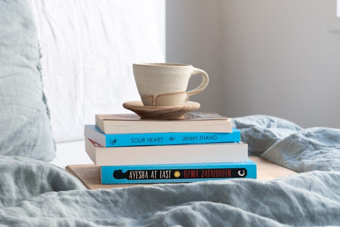 7 Books to Read from Ubud Writers and Readers Festival - Stack of 4 books on a bed with pale blue linen. Visible titles are Sour Heart by Jenny Zhang and Ayesha at Last by Uzma Jalaluddin.