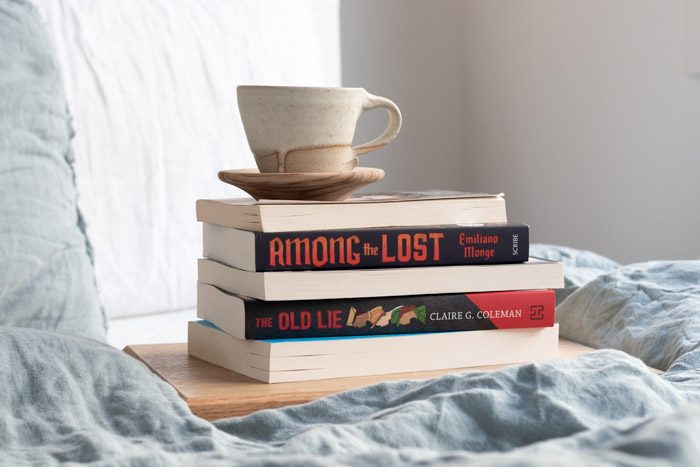 7 Books to Read from Ubud Writers and Readers Festival - Stack of 4 books on a bed with pale blue linen. Visible titles are Among the Lost by Emiliano Monge and The Old Lie by Claire G. Coleman.