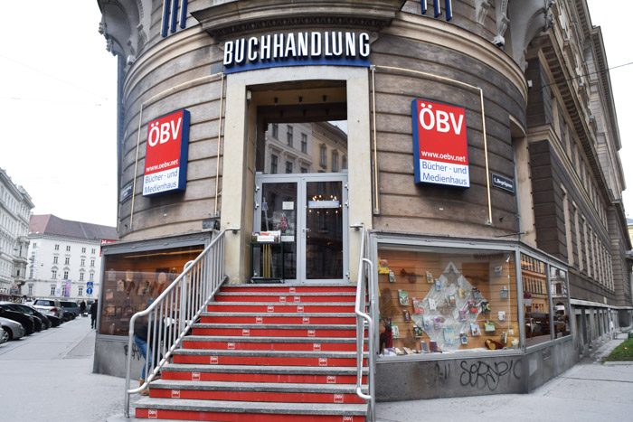 A rounded, period shopfront with the shop's name (Buchhandlung) above the entrance and two signs with the shop's initials (OBV) on either side of the entrance. Stairs lead up to the front door.