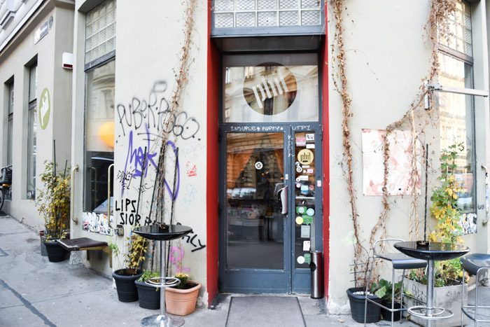 A white, rendered shopfront and blue trim door, surrounded by a few small potted plants and with graffiti on one wall.