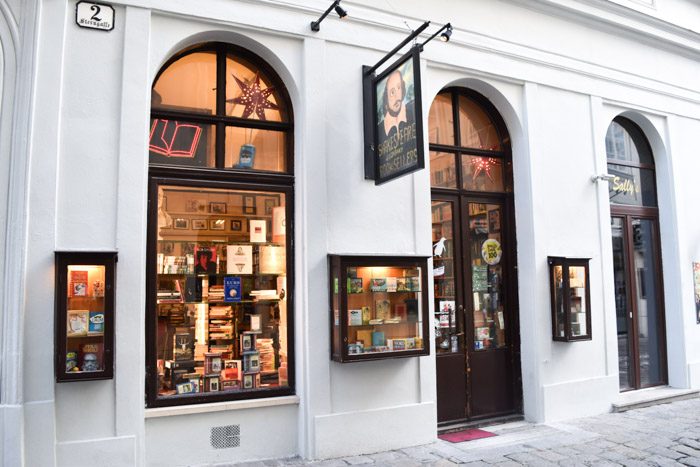 White rendered shopfront with arched windows showing the illuminated interior of Shakespeare and Company Booksellers.